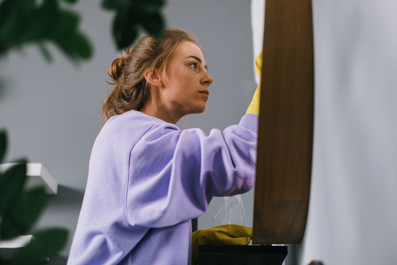 Side profile of a young focused woman wearing latex gloves, standing and cleaning a mirror in a bright room during the day